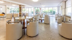 A spacious hotel lobby with round tables, cushioned seating, and large windows offering a view of the outdoors. - HOTEL NAICA (Rimini)