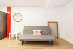 A modern living room with a grey sofa, a small white table, a red accent wall, a gold-framed mirror, and a framed picture.