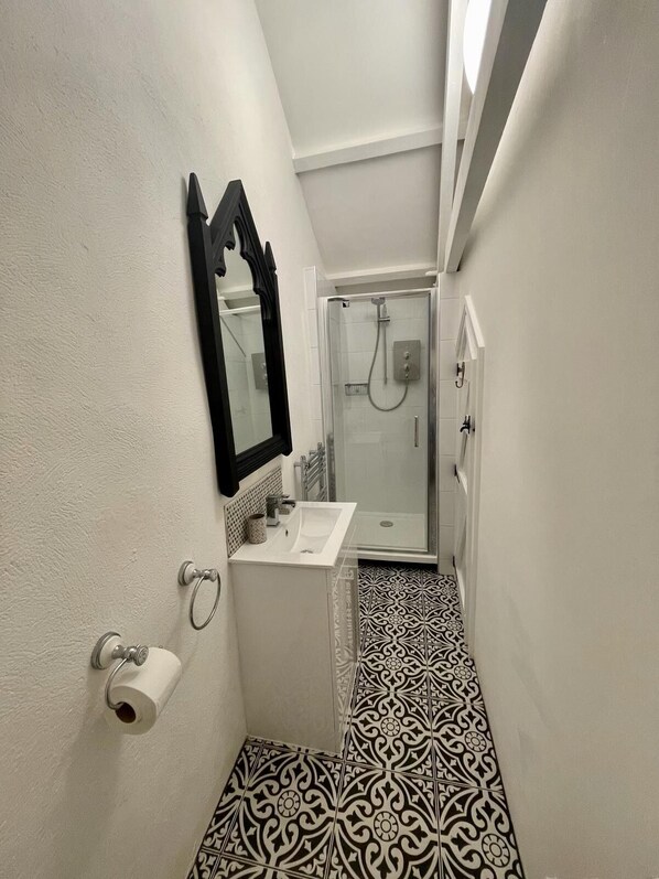 A narrow bathroom with a white sink, a toilet, and a shower. The floor has a black and white patterned tile.