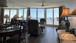 Ett rymligt rum med ett stort fönster som erbjuder utsikt över havet, en matplats med bord och stolar samt en sittgrupp med en soffa och fåtöljer.