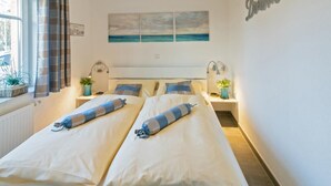 A double bed with matching pillows and a striped blanket, a bedside table with a lamp, a window with curtains, and a wall-mounted artwork. - Ferienwohnung Kleine Auszeit (Norddeich)