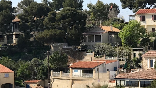 Un port de plaisance sur la côte, avec des bateaux amarrés, un petit bâtiment portant une enseigne, et des maisons en arrière-plan.