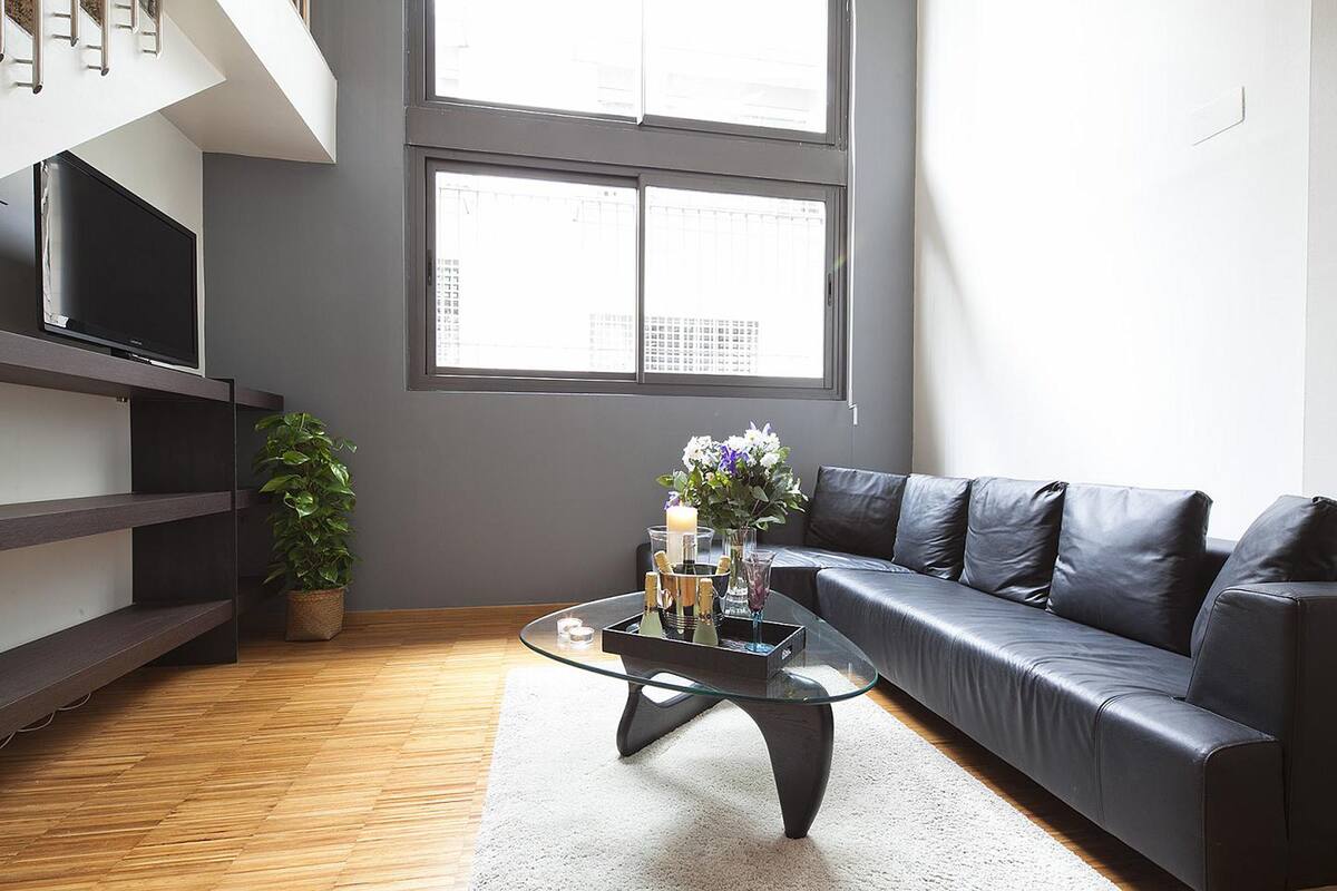 A modern living room with a glass coffee table, a black leather sofa, a TV on a shelf, and a large window with a city view.