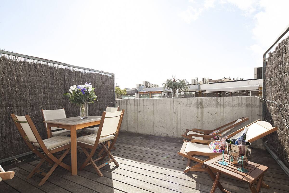 A rooftop terrace with a wooden table and chairs, a potted plant, and a bottle of champagne.
