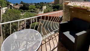 Un balcon doté d’une table ronde en métal et de meubles en osier, offrant une vue sur un paysage côtier avec des maisons et des arbres.