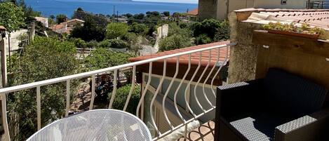A balcony with a round metal table and wicker furniture, overlooking a coastal landscape with houses and trees.