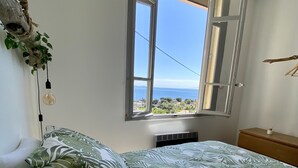 Une chambre à coucher avec un lit recouvert d’une courtepointe ornée d’un motif de feuilles tropicales, une fenêtre avec un volet entrouvert et une vue sur la mer.