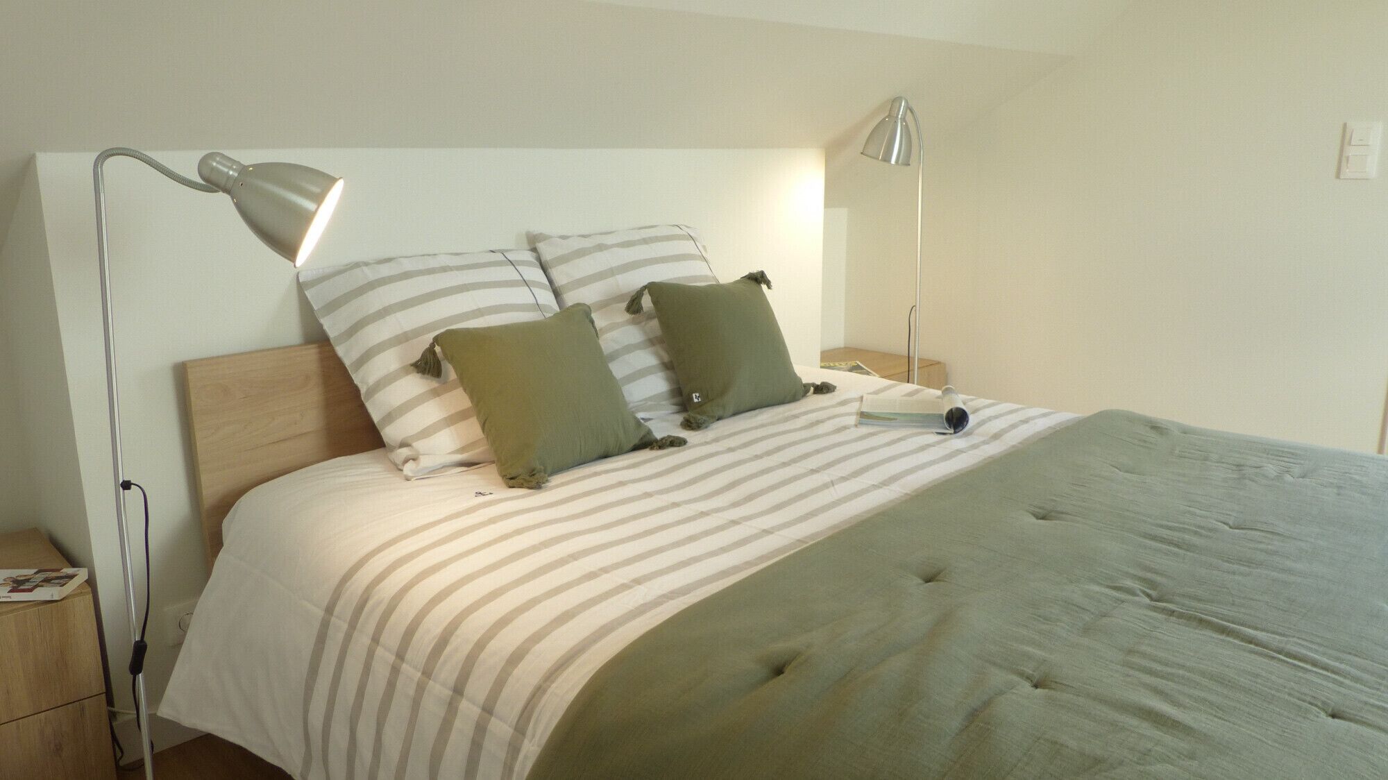 A neatly made bed with striped bedding, two pillows, and a green throw blanket. There are two bedside tables with lamps, and a small bedside table with a book and a cup.