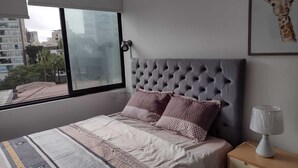 A bedroom with a tufted headboard, a large window, and a nightstand with a lamp.