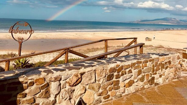 A beach with a stone wall and a wooden railing, a sign with the word "Calhemo", and a rainbow in the sky. - 207 - Apartamento Tarifa Sun (Tarifa)