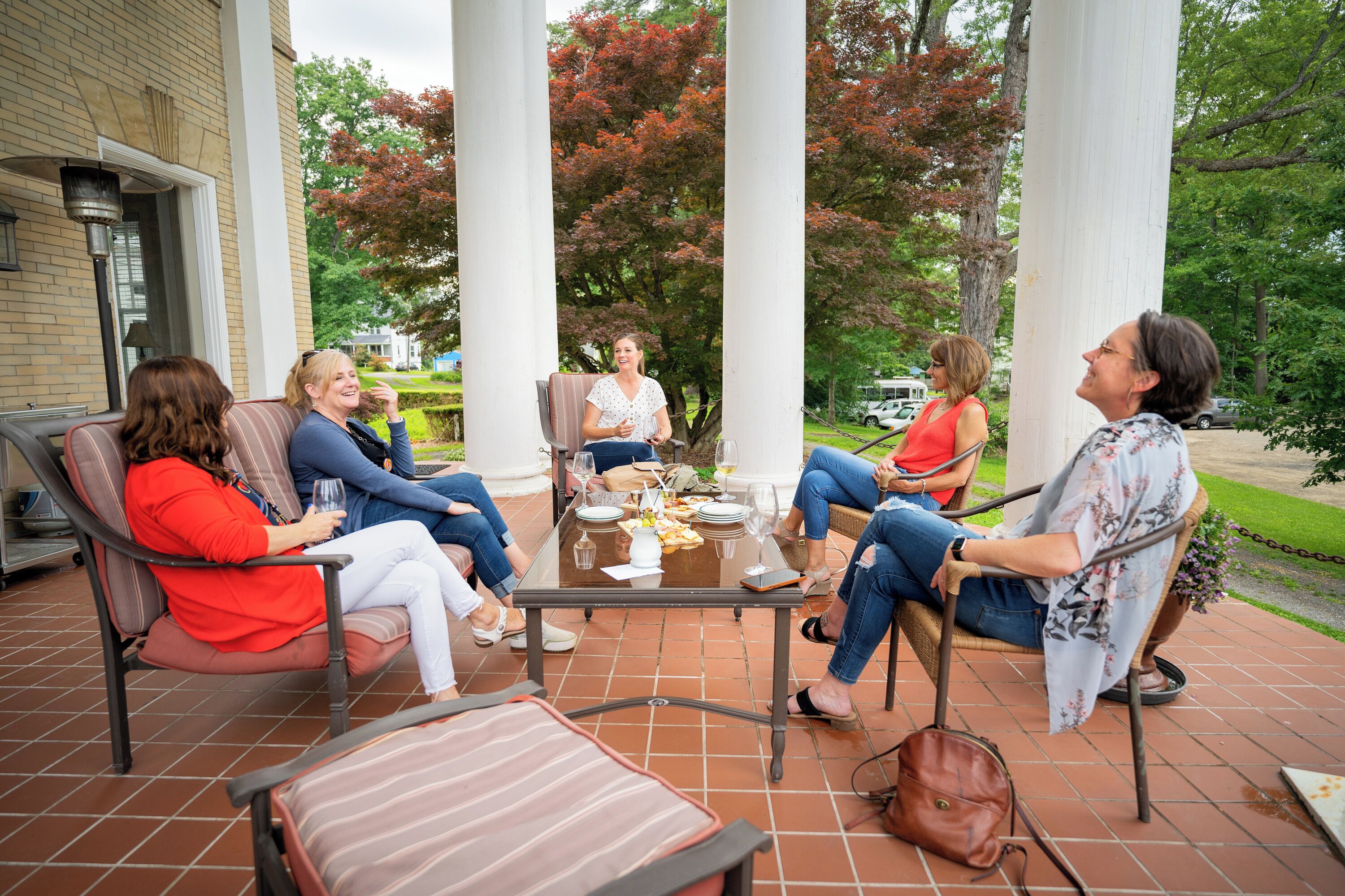 Un groupe de personnes est assis autour d’une table sur une terrasse dont le sol est pavé de briques, et l’ensemble est entouré de colonnes blanches et d’arbres aux feuilles d’automne.