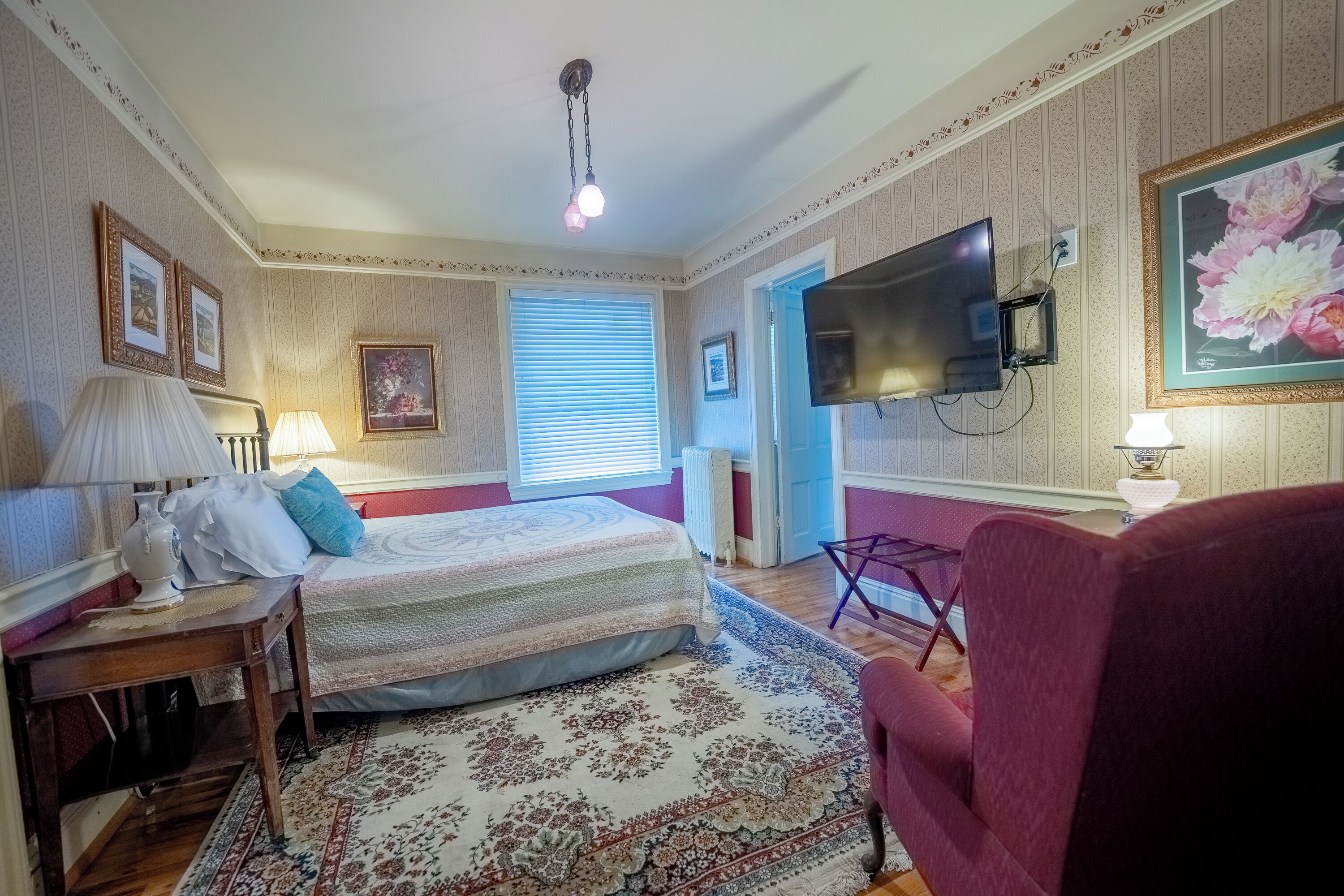 A bedroom with a bed, a TV mounted on the wall, a red armchair, and a large window with blinds.