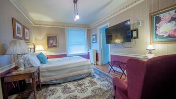 A bedroom with a bed, a TV mounted on the wall, a red armchair, and a large window with blinds.