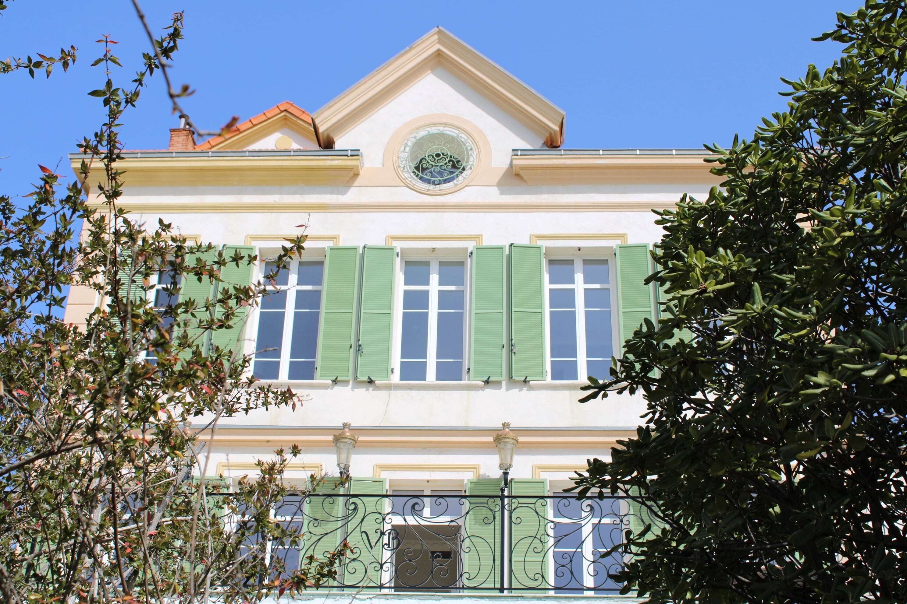 A two-story building with green shutters, a central circular window, and a decorative balcony railing.