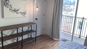 Un dormitorio con piso de madera, una cama con ropa blanca, un escritorio con estantes, un cuadro en la pared y vistas a través de una ventana.