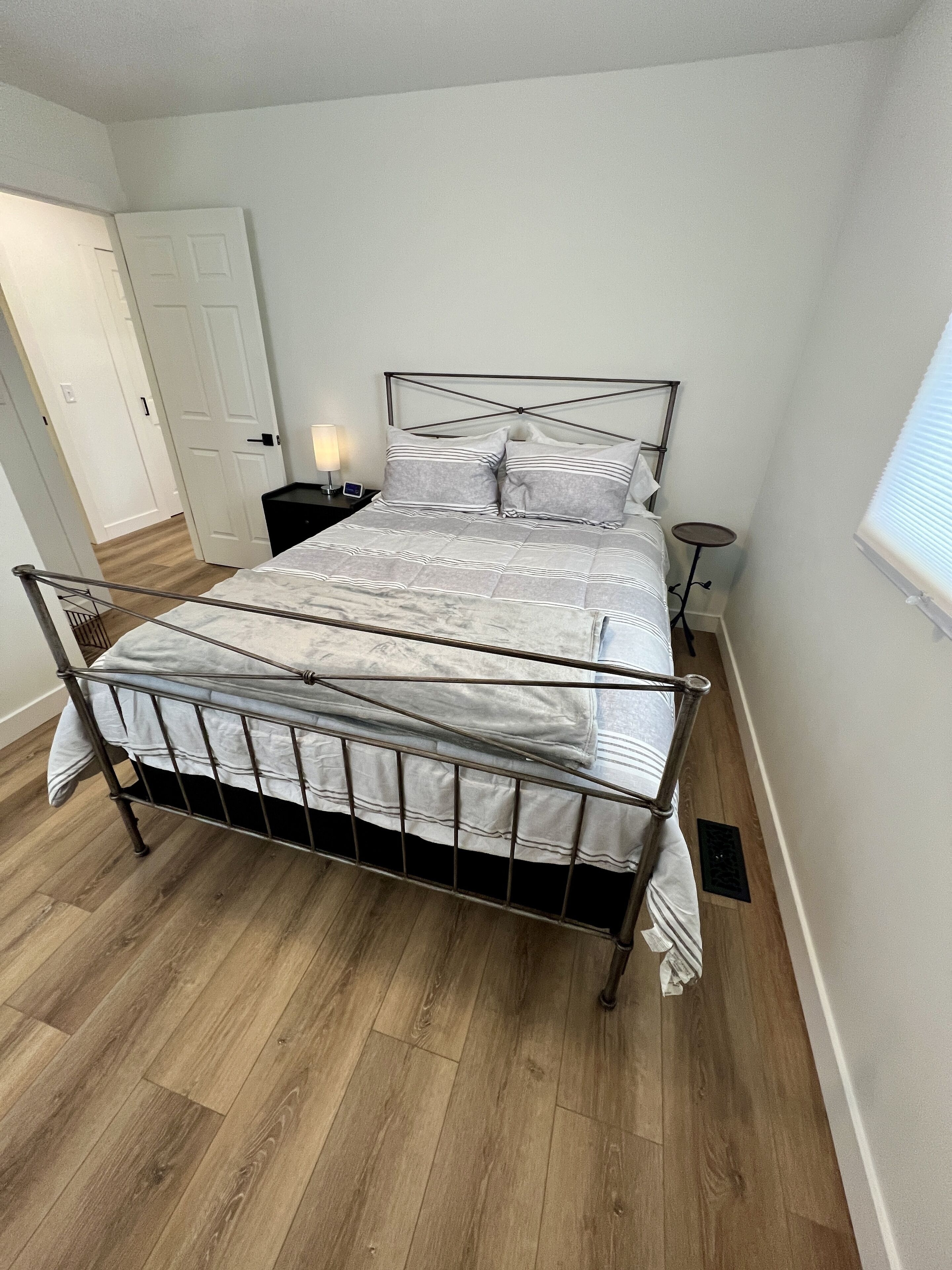 A bedroom with a metal bed frame, a white wall, a wooden floor, a bedside table with a lamp, and a window with blinds.