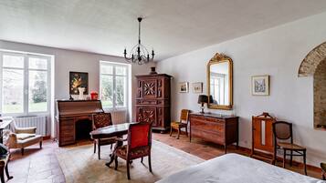 Une chambre spacieuse avec un grand lit, une commode en bois, un bureau et une chaise. On y trouve une cheminée, un lustre et un grand miroir au mur.