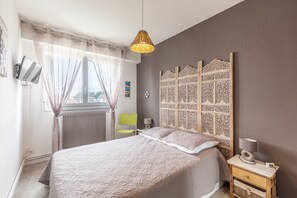 Un dormitorio con una cama grande, una mesita de noche con lámpara, un cabecero decorativo y una ventana con cortinas translúcidas.