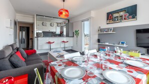 Een moderne woonkamer met een grijze bank, een eettafel voor zes personen gedekt met rood en wit servies, een keuken op de achtergrond en een televisie aan de rechter muur.