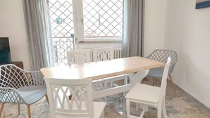 A dining area with a wooden table and white chairs, a TV on the left, and a window with a metal grid. - Sunnyhome- Ostia Lido sea view | 2 bathrooms (Lido di Ostia)