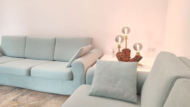 A light blue sofa with a cushion, a small side table with a decorative item, and a white wall. - Sunnyhome- Ostia Lido sea view | 2 bathrooms (Lido di Ostia)