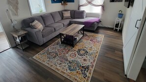 Una sala de estar moderna con un sofá seccional gris, una mesa de centro de madera y una alfombra colorida. Hay dos mesas auxiliares, una con una lámpara y un adorno. Se ve una ventana con cortinas translúcidas y vistas al exterior.