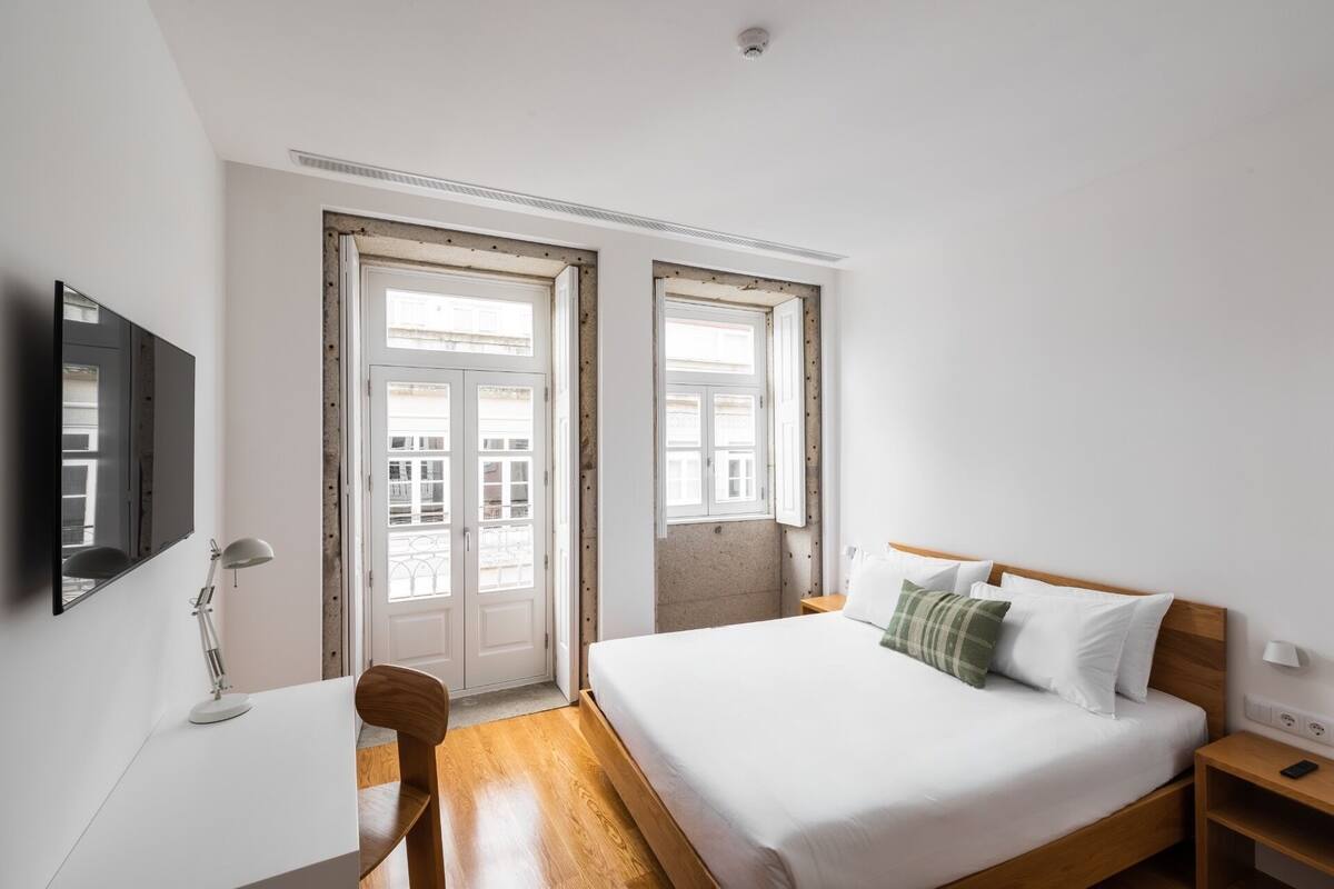 Un dormitorio con cama, escritorio, silla, televisor y dos ventanas con vista a edificios.