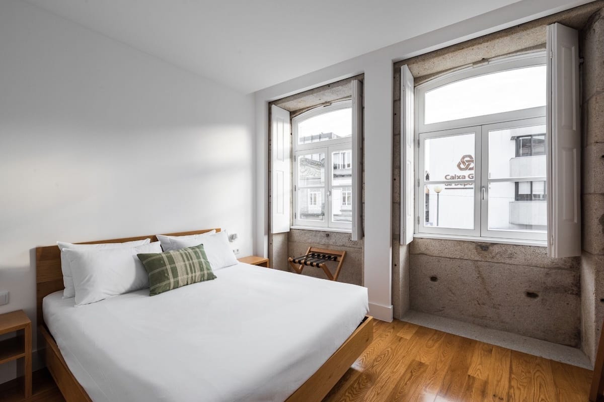 Un dormitorio con una cama grande, cabecera de madera, dos ventanas, un banco y vistas a edificios.