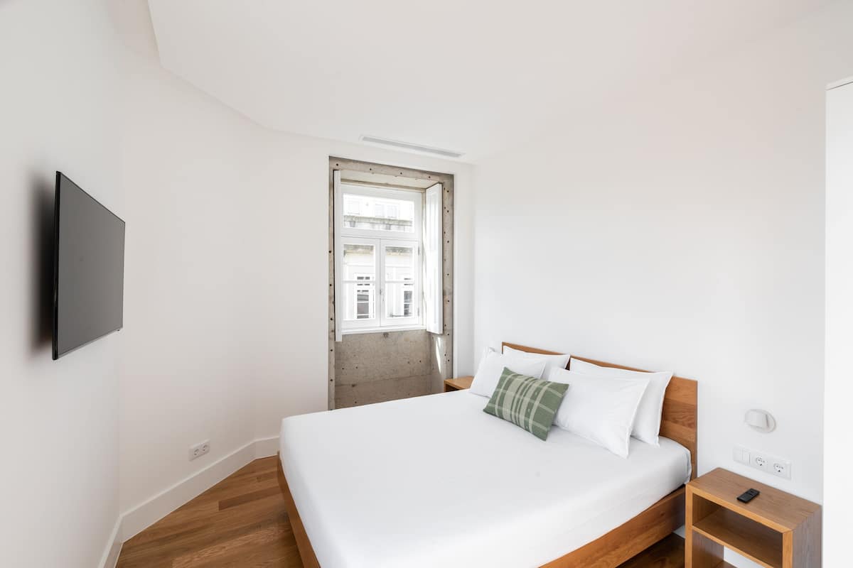 Un dormitorio con cama, televisor montado en la pared, mesita de noche de madera y ventana con vistas a edificios.