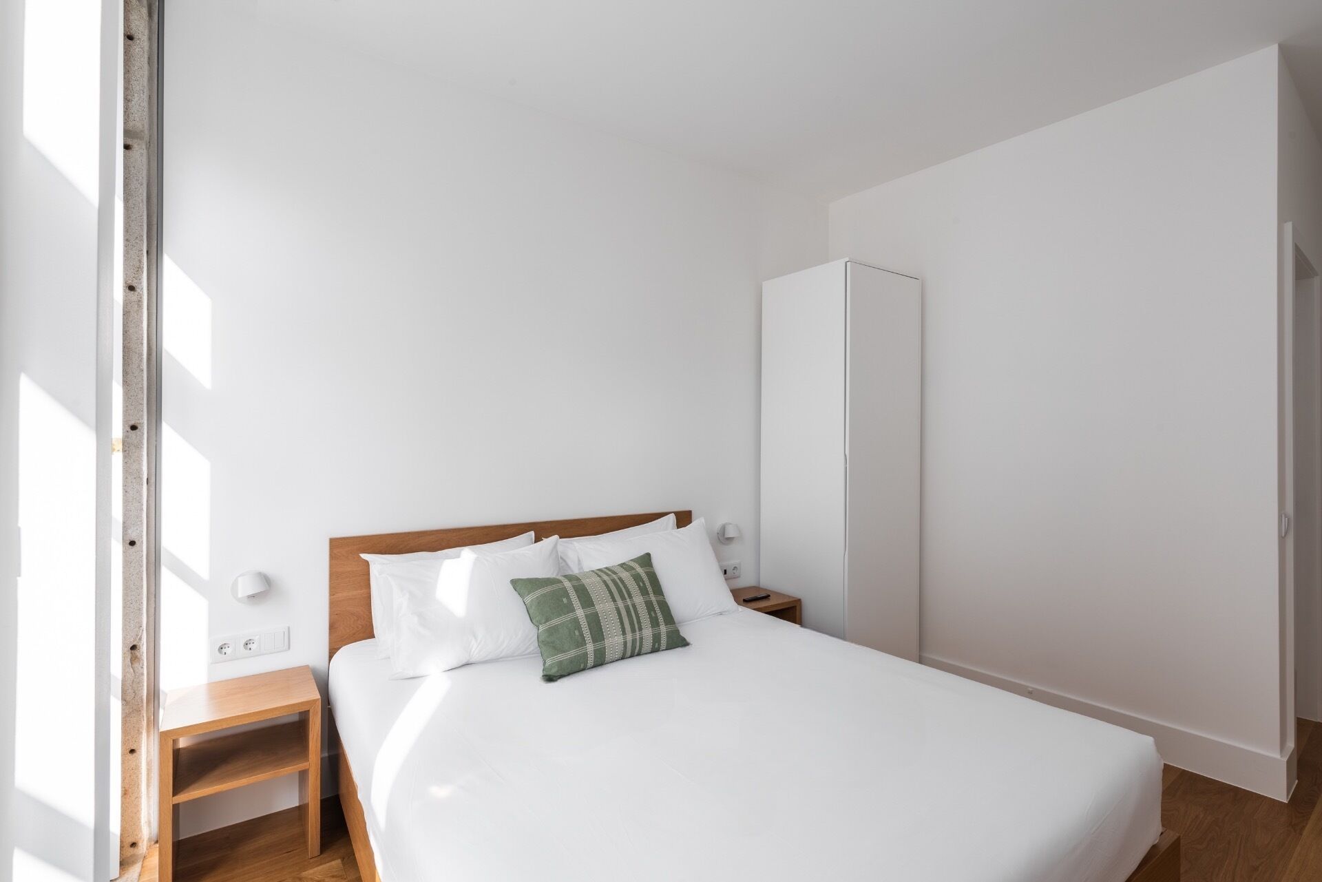 Een minimalistische slaapkamer met een houten hoofdbord, wit beddengoed, een groen kussen, een nachtkastje en een kledingkast.
