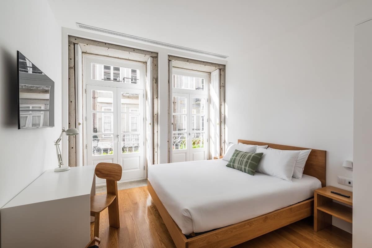 Un dormitorio con una cama de madera, ropa de cama blanca, un escritorio con lámpara y una silla.