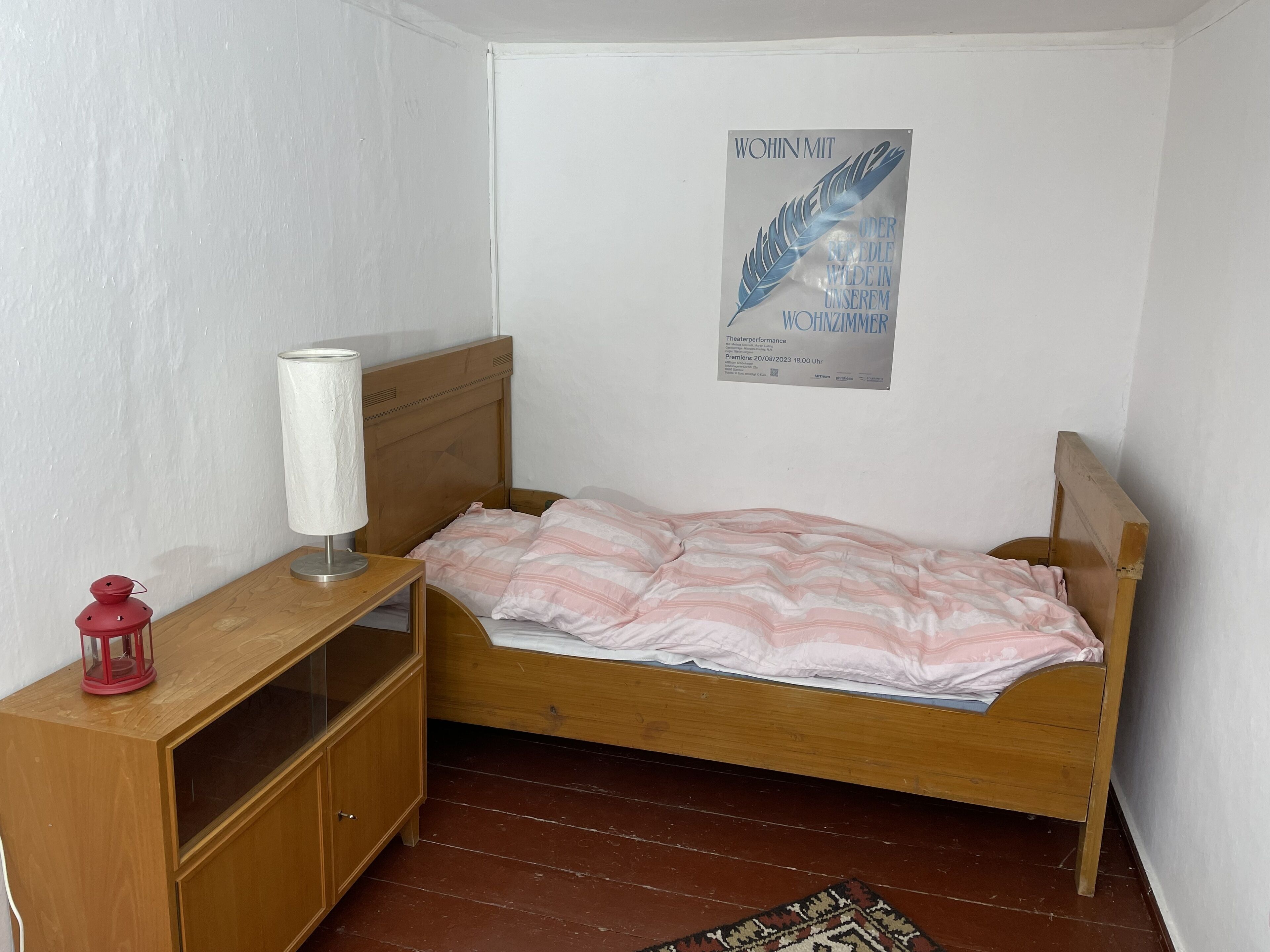 A small, neatly arranged room with a wooden bed, a bedside table with a lamp, and a poster on the wall.