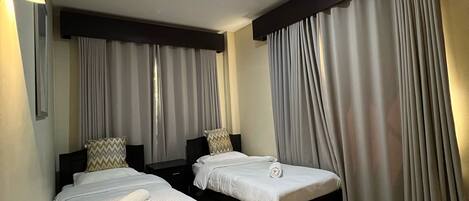 A hotel room with two single beds, each with a pillow and a rolled towel. The room has a large window with curtains, a ceiling light, and a dark-colored headboard.