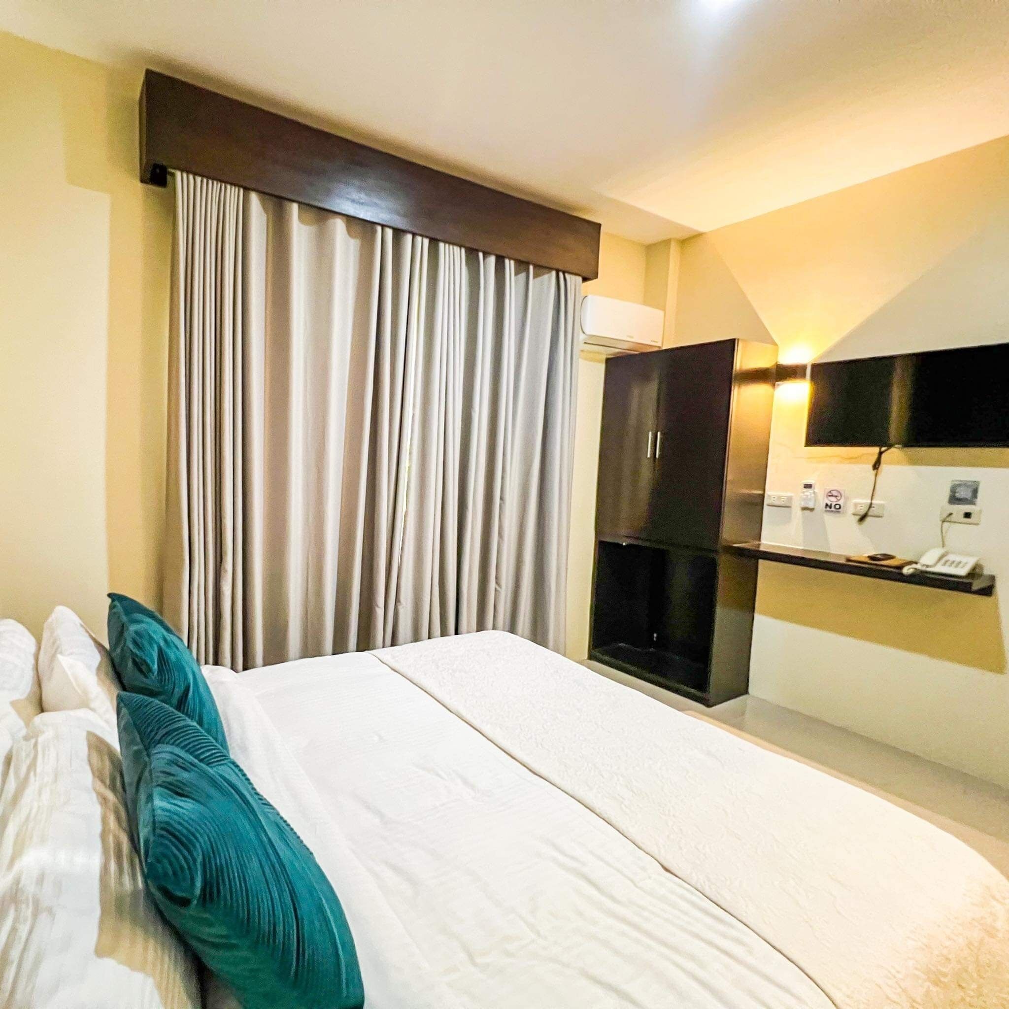 A hotel room with a bed, curtains, a wardrobe, a desk with a phone, and a wall-mounted TV.
