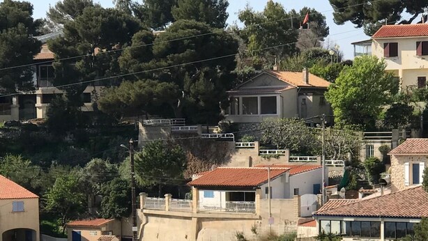 Un puerto costero con barcos amarrados, un pequeño edificio con un letrero y casas al fondo.