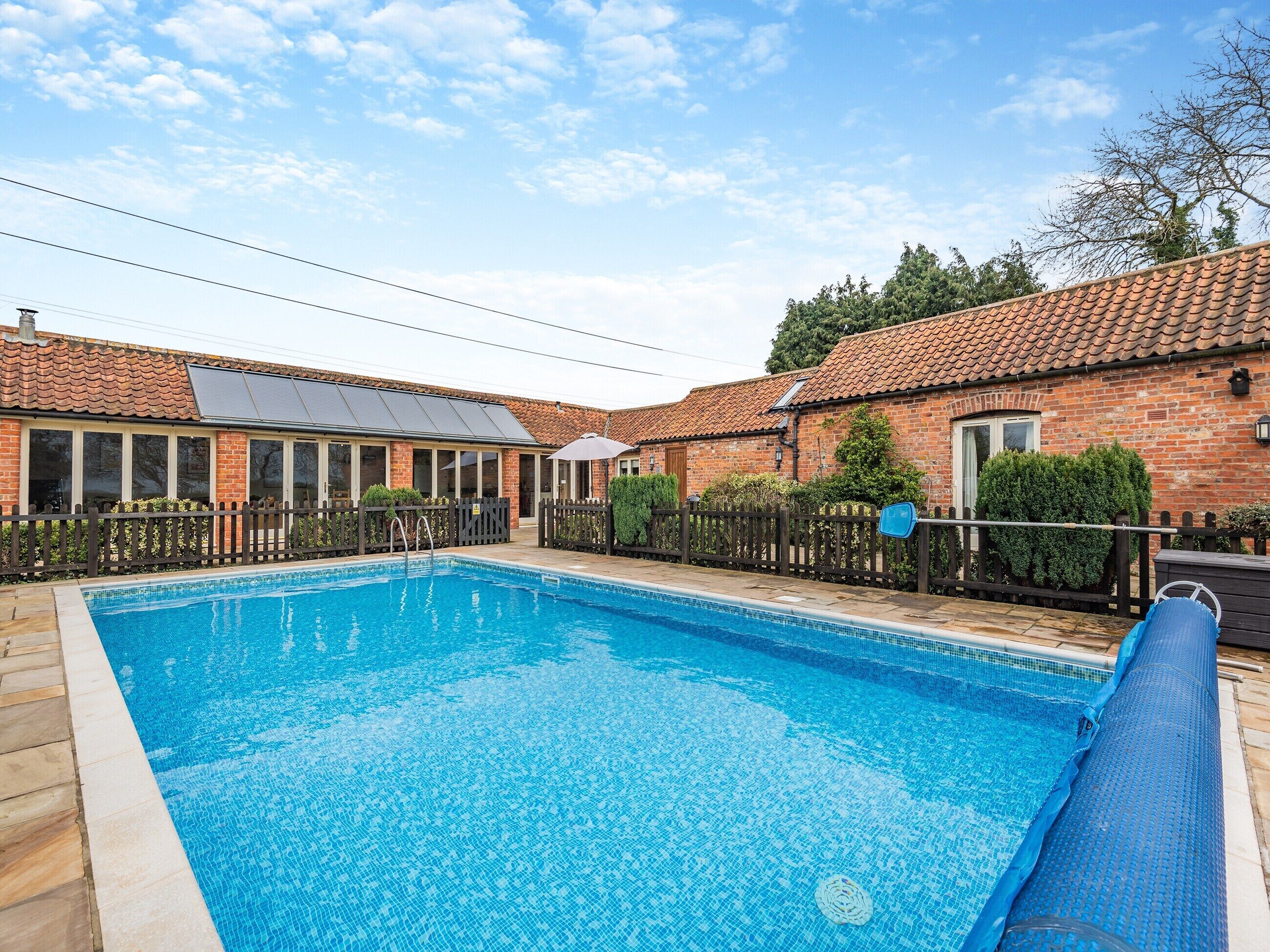A swimming pool with a tiled deck, surrounded by a brick building with a tiled roof and a fenced patio area.