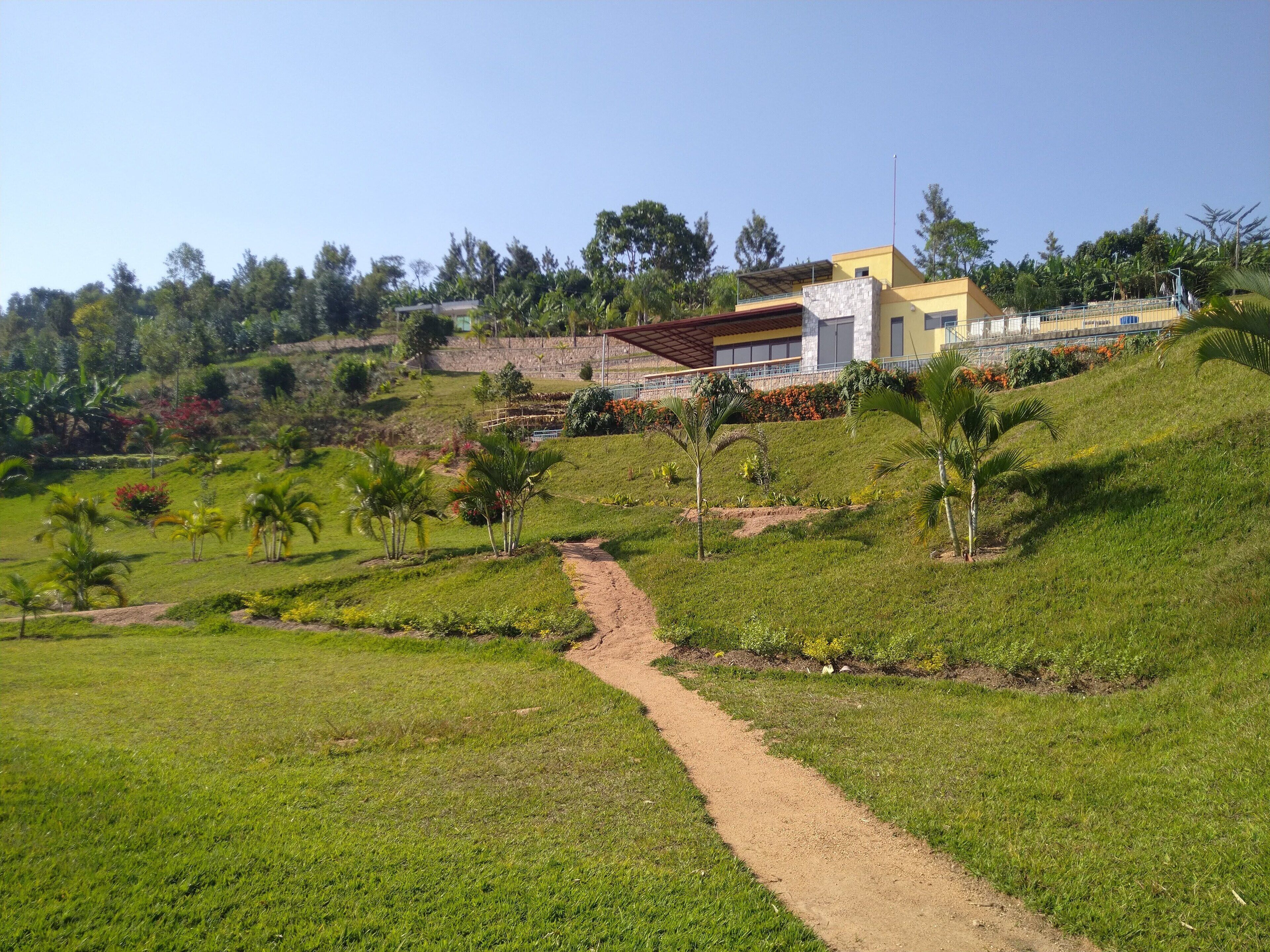 Um terreno em encosta com um caminho de terra que leva a uma casa moderna com fachada amarela e telhado vermelho.