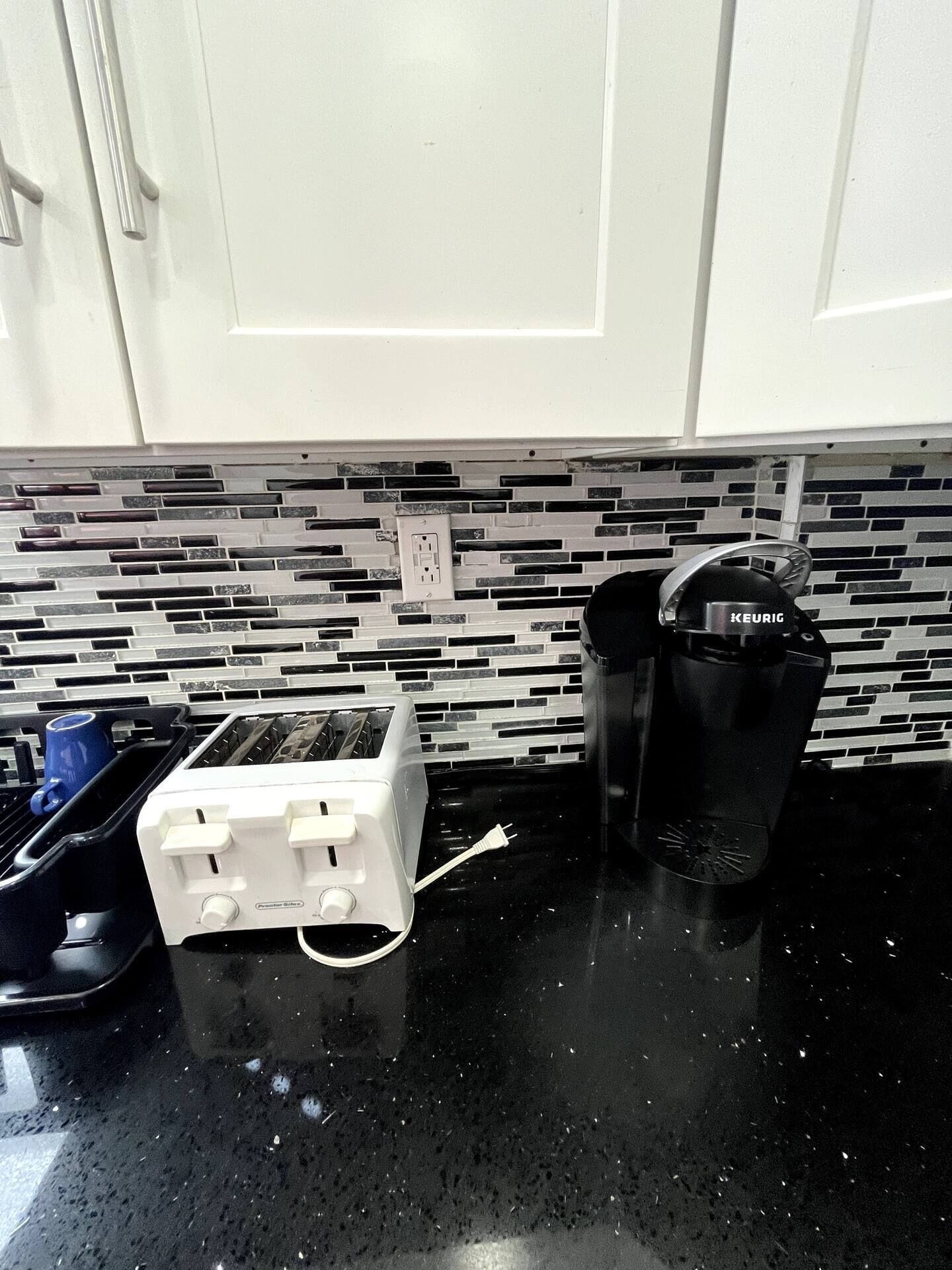 Et køkken med hvide skabe, en sort bordplade, en brødrister, en Keurig kaffemaskine og et flisebeklædt bagpanel.