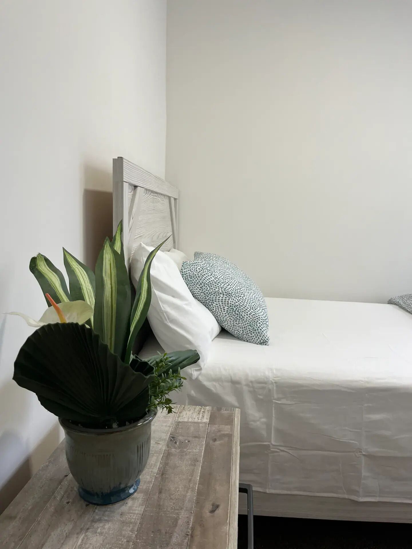 A bedroom with a bed, a bedside table with a potted plant, and a headboard against a plain wall.