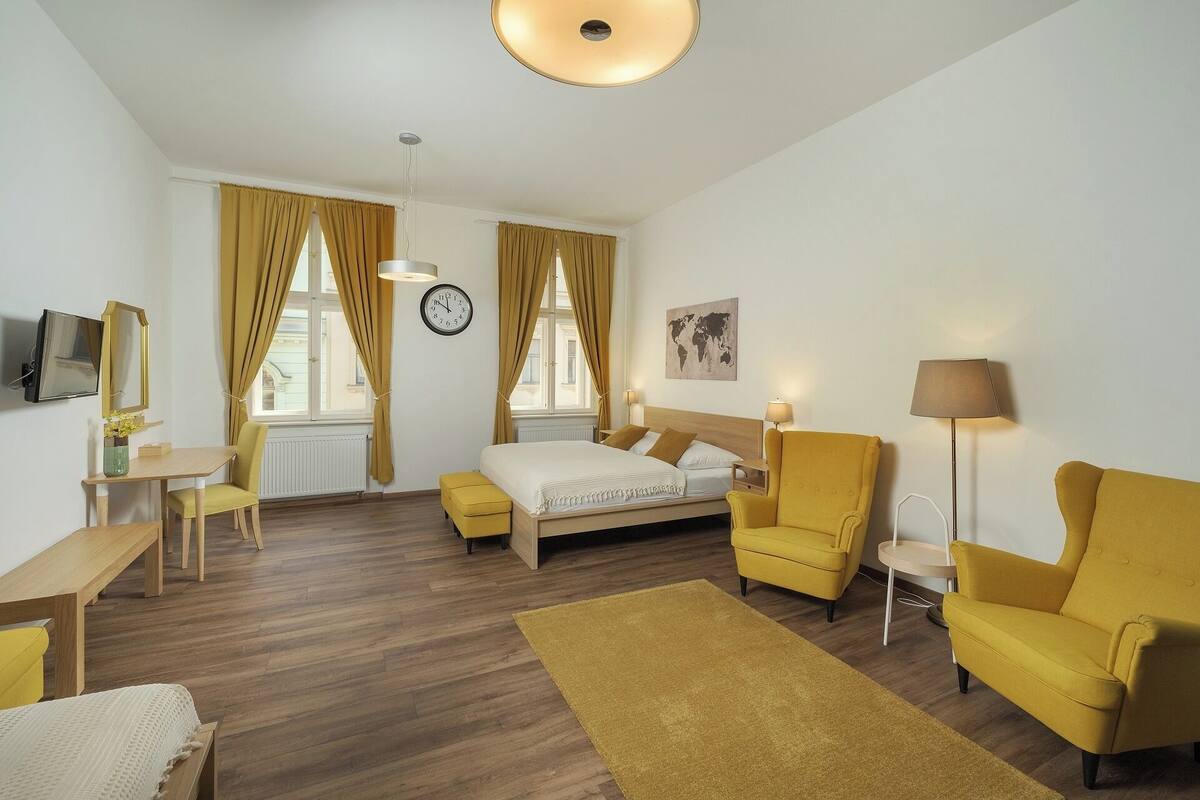 Habitación de hotel con cama, dos sillones amarillos, escritorio, televisor, mapa mundial en la pared y ventana grande con cortinas.