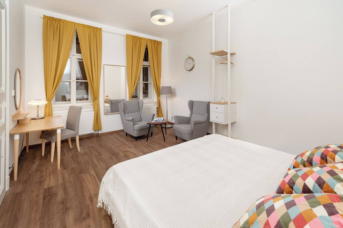 Habitación de hotel con cama, escritorio, dos sillas, lámpara, reloj y armario.