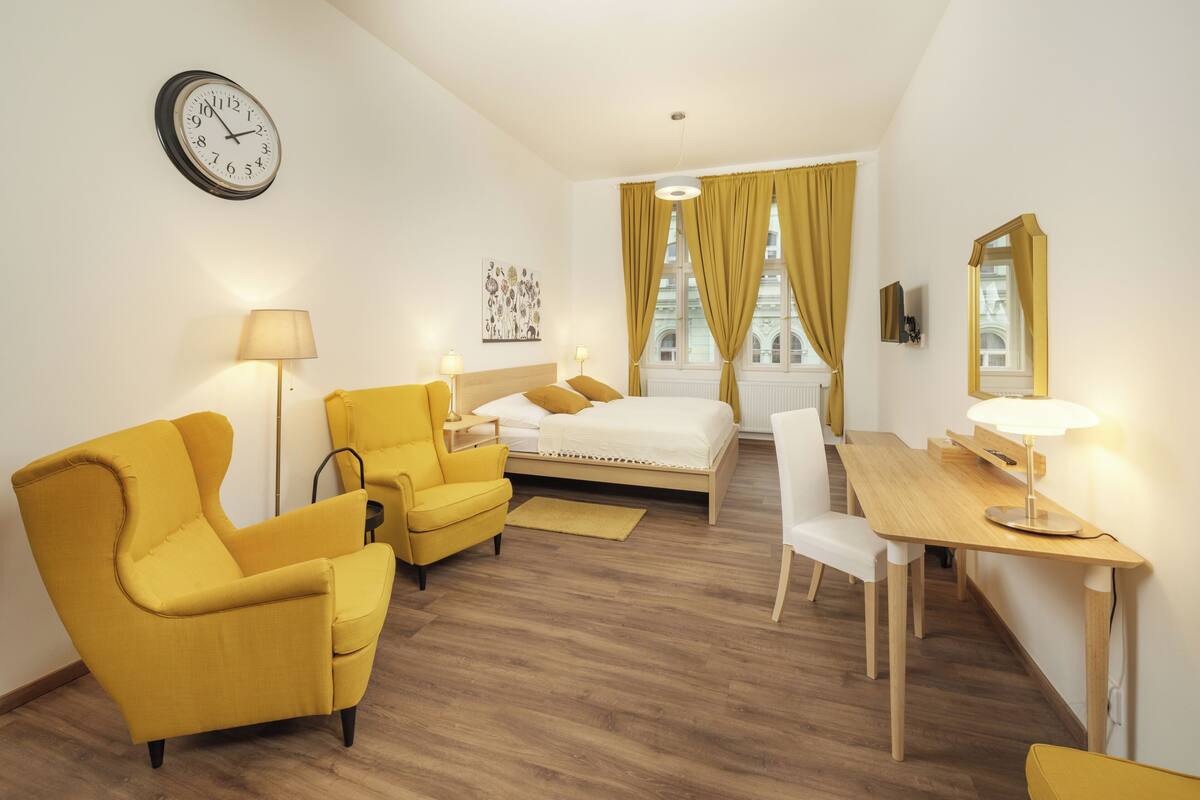Una habitación de hotel con una cama, un escritorio, dos sillones amarillos, una silla blanca, un reloj y un cuadro en la pared.