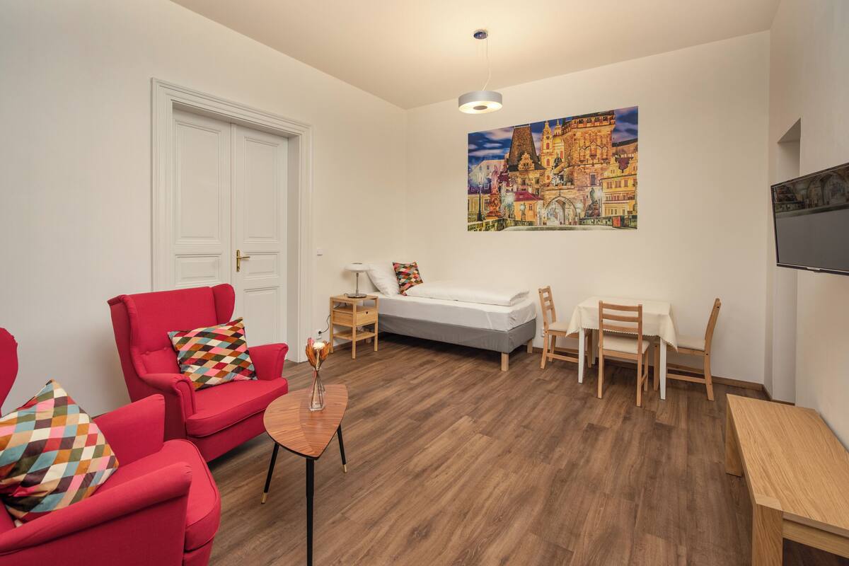 Una sala de estar moderna con una cama, sillas rojas, una mesita y un cuadro en la pared.