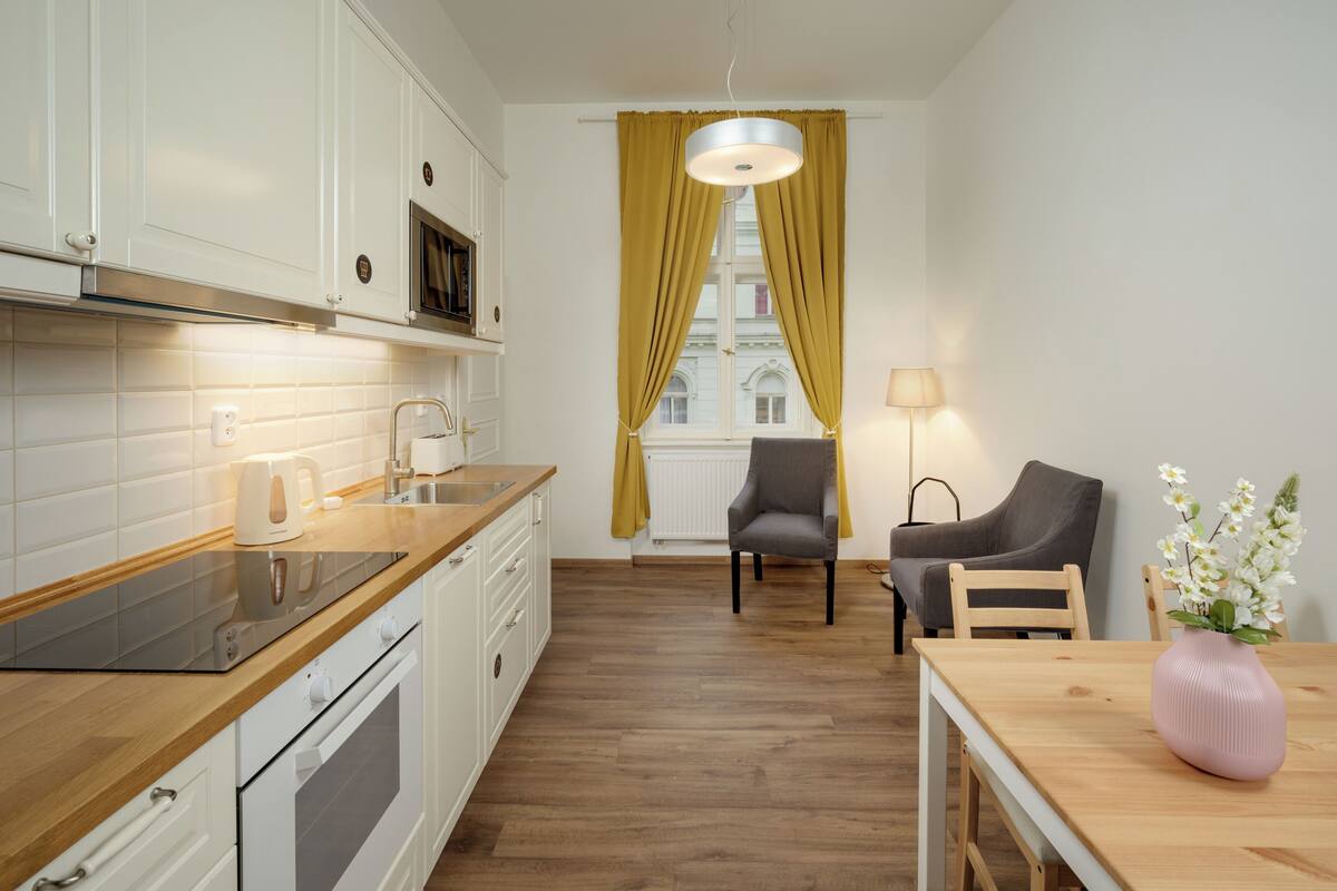 Una cocina moderna con armarios blancos, encimera de madera y horno empotrado. Hay una zona de comedor con una mesa y sillas de madera, y un florero con flores sobre la mesa.