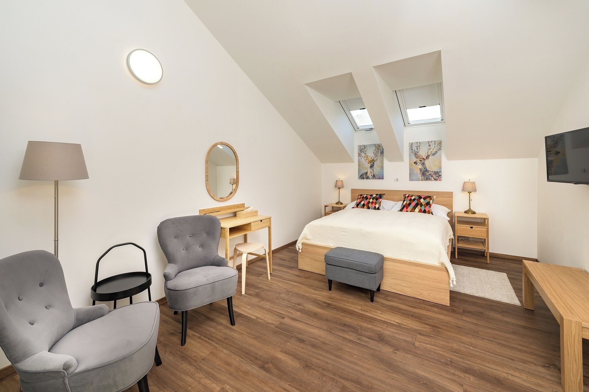 A modern bedroom with a bed, two chairs, a desk, a mirror, and a TV.