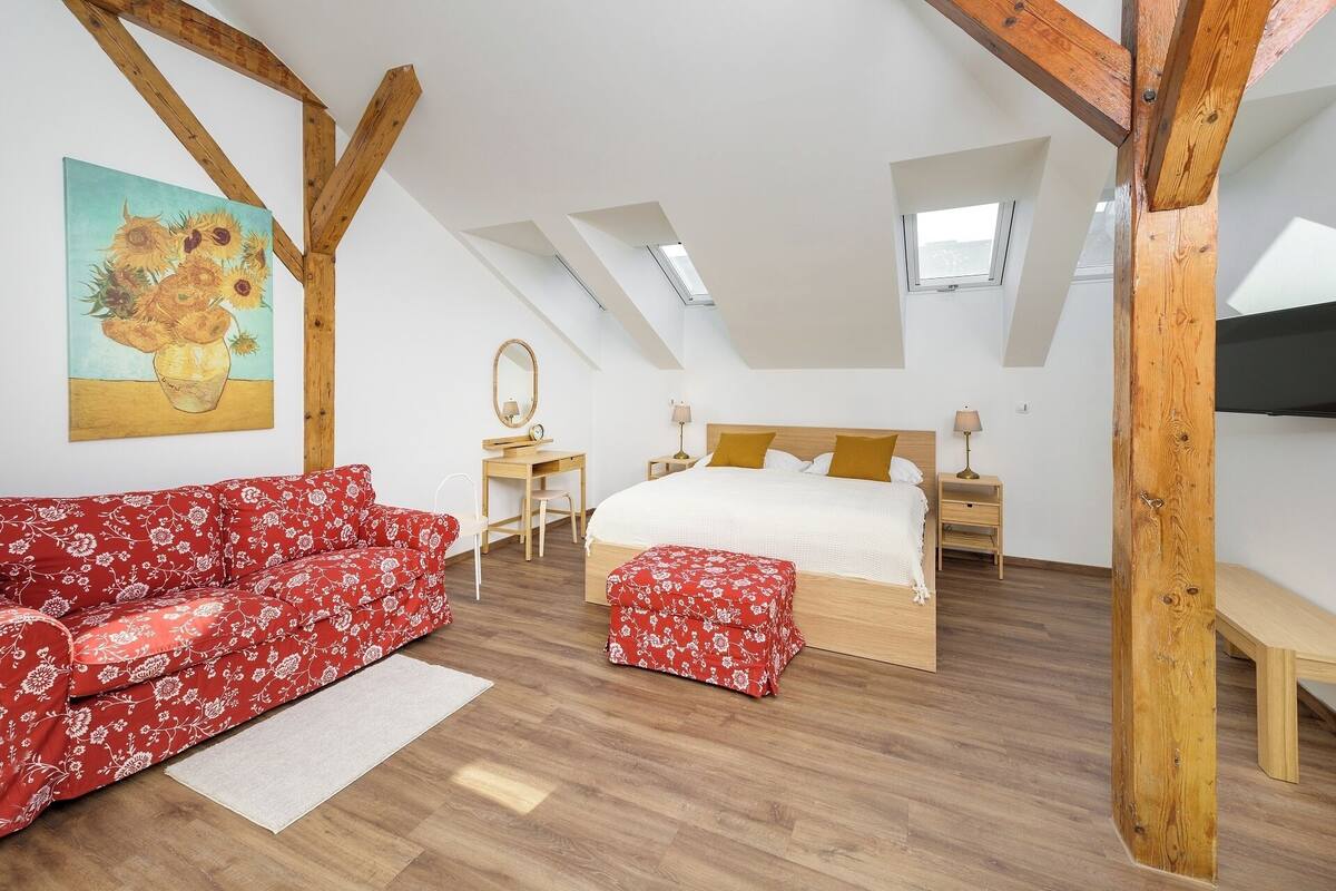 Un dormitorio con cama, un sofá floral rojo, una mesita y un cuadro de girasoles.