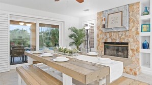 A dining area with a wooden table set for four, a stone fireplace, and a ceiling fan.