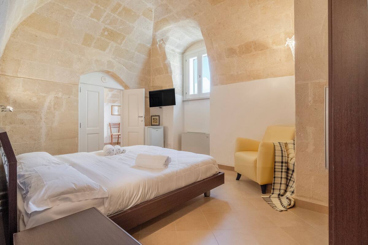 A bedroom with a stone ceiling, a bed with white linens, a yellow armchair, and a small table.