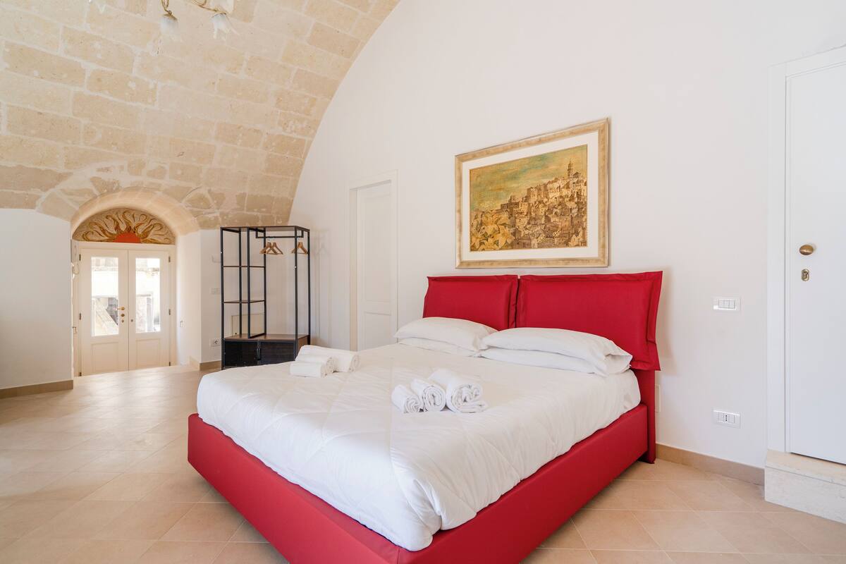 A bedroom with a red bed, white bedding, a framed picture on the wall, and a stone wall.