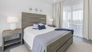 Un dormitorio moderno con cabecera de madera, una cama grande con colcha estampada, dos mesitas de noche con lámparas y vistas a la ciudad a través de cortinas translúcidas.
